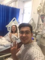 Living kidney donor Henry and his sister and recipient. The two are in hospital and are both wearing hospital gowns. Henry is taking a selfie of them both smiling and waving for the camera.